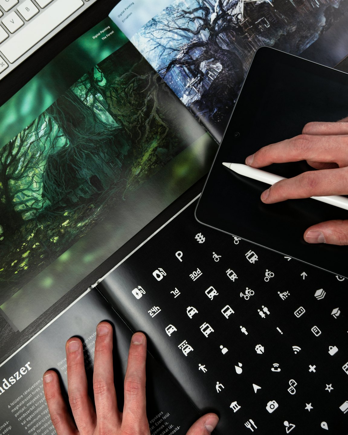 Hände benutzen einen Stift auf einem Tablet neben einer Zeitschrift und Tastatur.